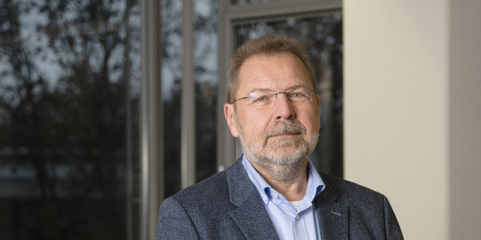 Portraitfoto von Prof. Dr. Thomas Goll, im Hintergrund: Unscharf abgebildetes Büro, außen Vegetation.