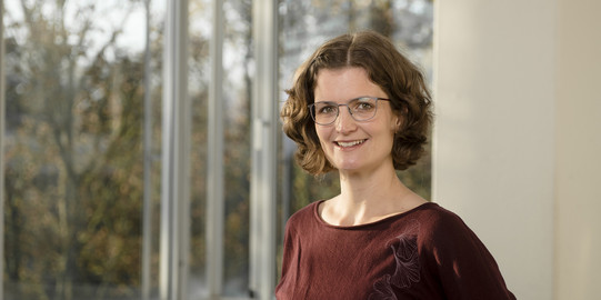 Portraitfoto von Dörte Kanschik, im Hintergrund: Unscharf abgebildetes Büro, außen Vegetation.
