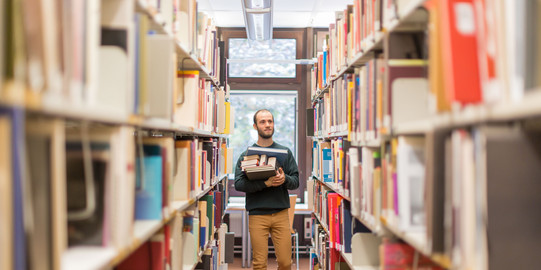 Studierender läuft an den Bücherregalen in der Bibliothek entlang.